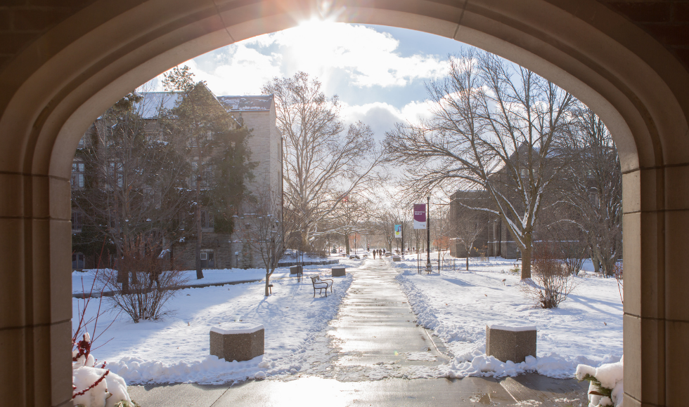 A photo of the archway on McMaster's campus. There is snow on the ground and sunshine is peeking through the top of the archway.