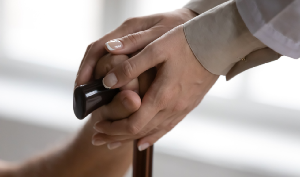A photo of a person's hand resting on a cane. Another set of hands are enclosed around the hand.