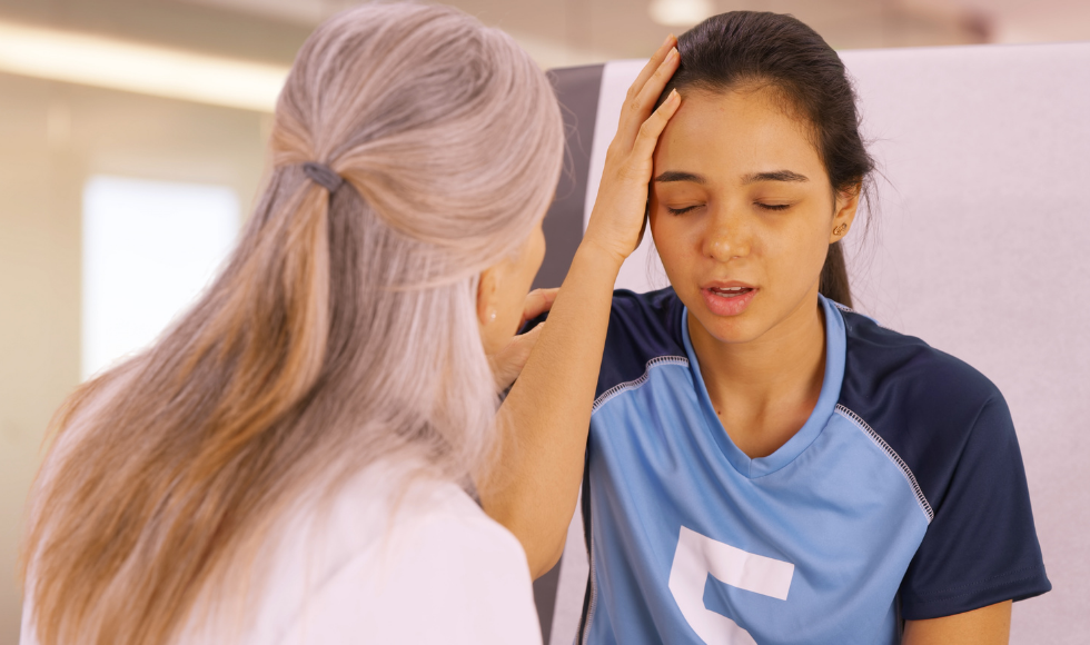 A young woman in a sports uniform touches her head while a person in a lab coat talks to them.
