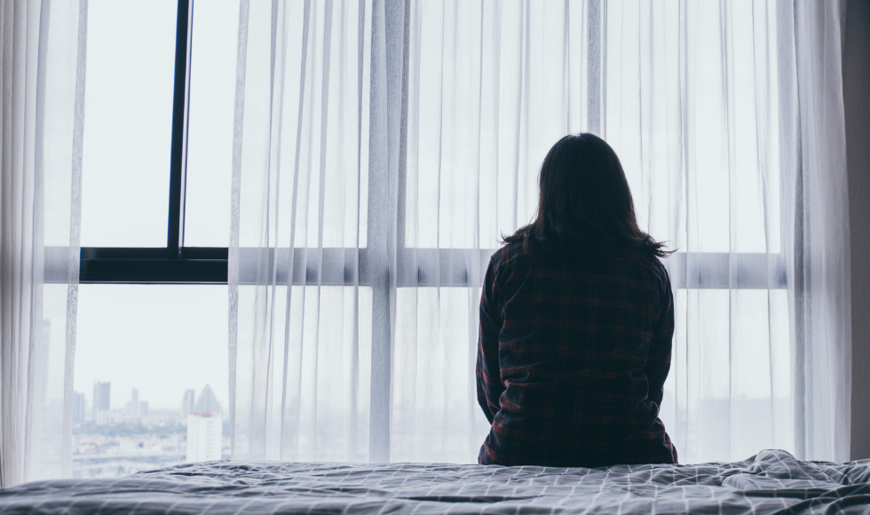 The silhouette of someone with long hair looking sitting on a bed and looking out the window