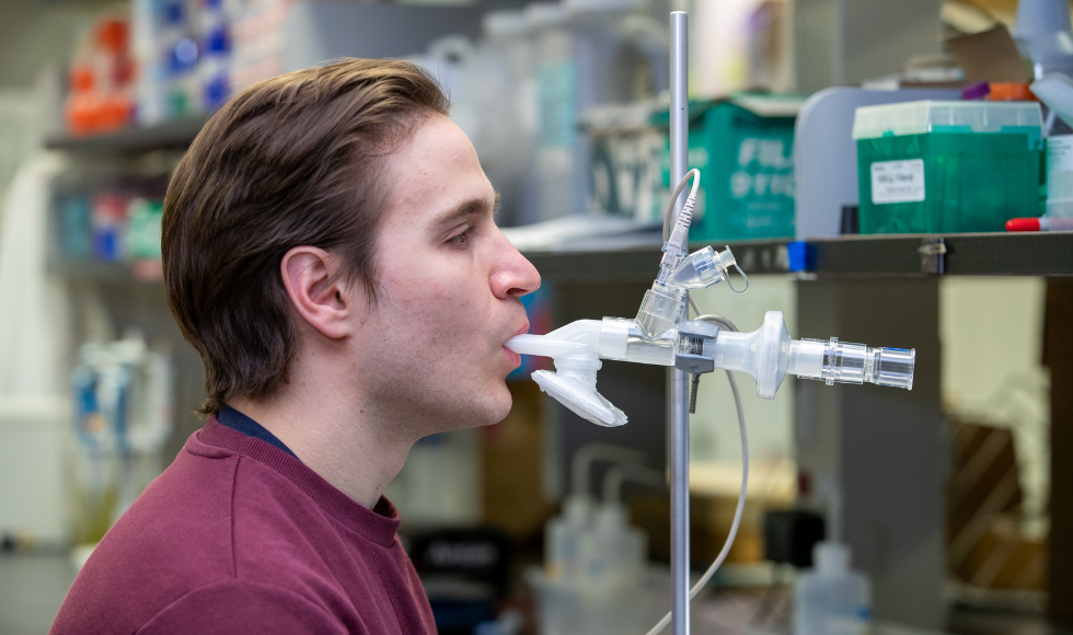 Man in red shirt inhales vaccine through an inhaled delivery system in a lab setting.