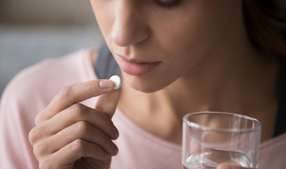 A photo of a woman's hands holding a pill and a glass of water. The woman is about to put the pill in her mouth.