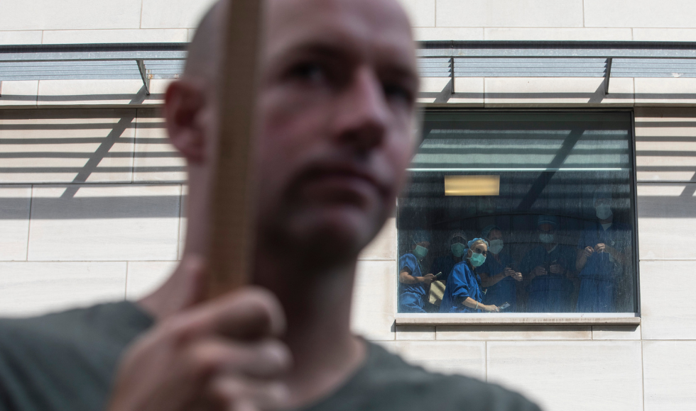 Man carrying a picket sign in the foreground; in the background is a wall with masked people in scrubs looking out a window