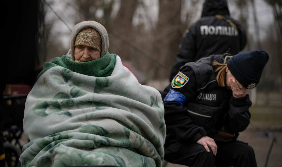 An older woman is wrapped in a blanket. Beside her is a Ukrainian police officer visibly upset and holding her head in her hand.
