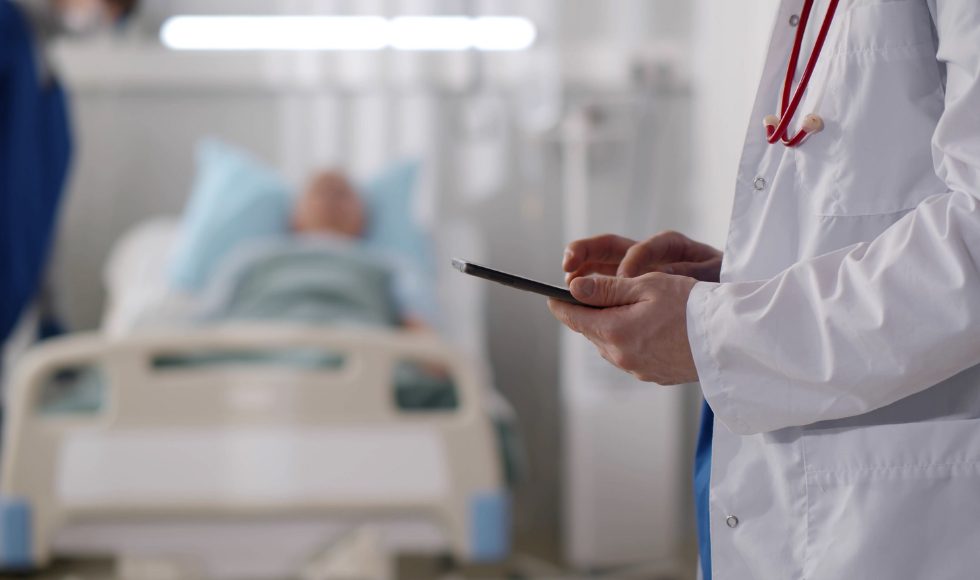 The hands of someone wearing a lab coat with a stethoscope hanging over their shoulders and holding a tablet. A patient lying in a hospital bed can be seen out of focus in the background.