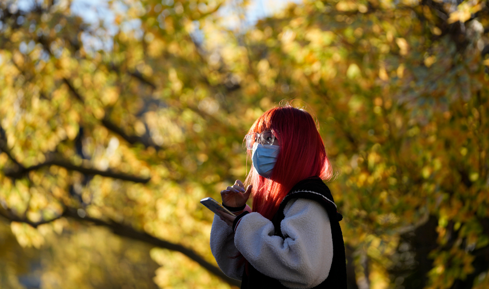A masked woman holding a phone standing in front of greenery