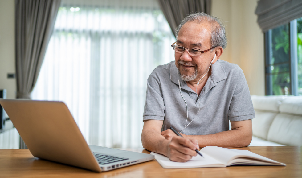 Older man with headphones sitting at a table holding a pen over a notebook and smiling into a laptop screen.