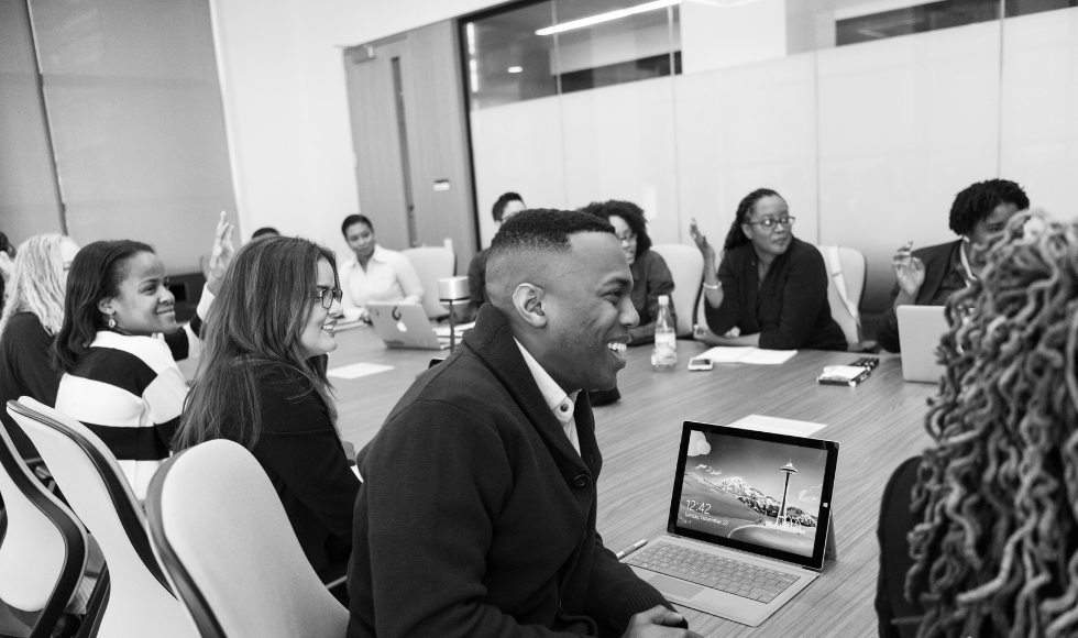A racially mixed group sitting around a conference table, smiling. A smiling Black man is closest to the camera.