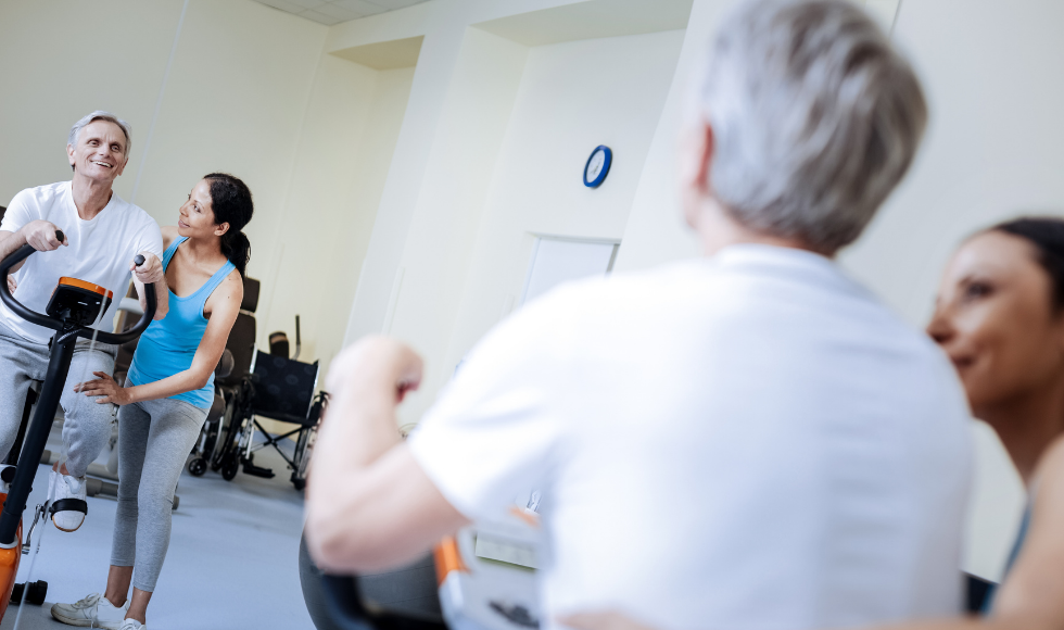 An older adult uses a stationary bike, assisted or encouraged by another person.