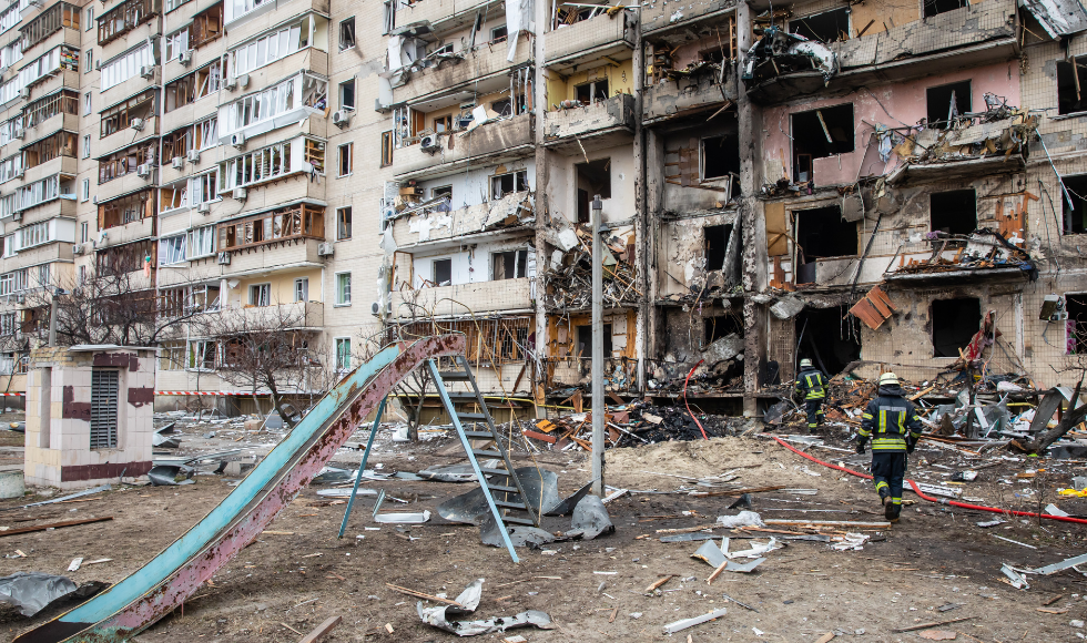 A person in an emergency worker's vest seen from behind walking through a destroyed playground toward burned and bombed-out buildings in Kyiv.