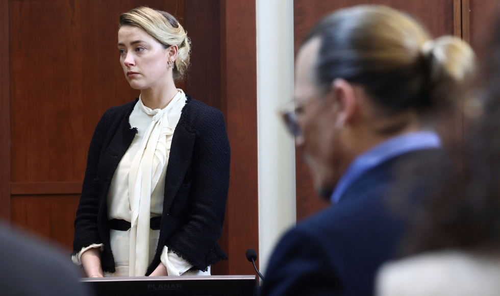 Amber Heard standing in a courtroom. Johnny Depp is seated and out of focus in the foreground