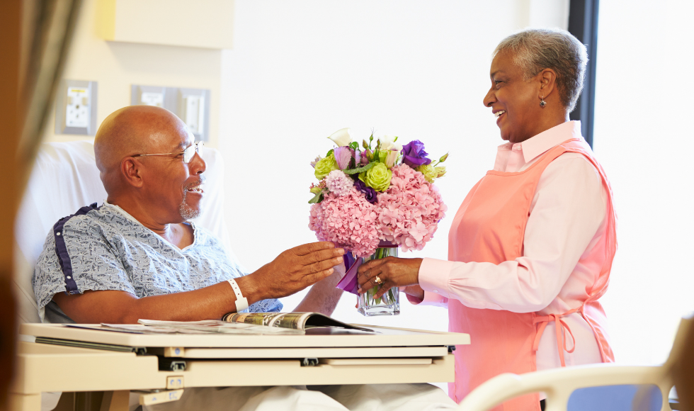 An older adult in a hospital bed smiles as he is handed flowers by a hospital worker or caregiver.