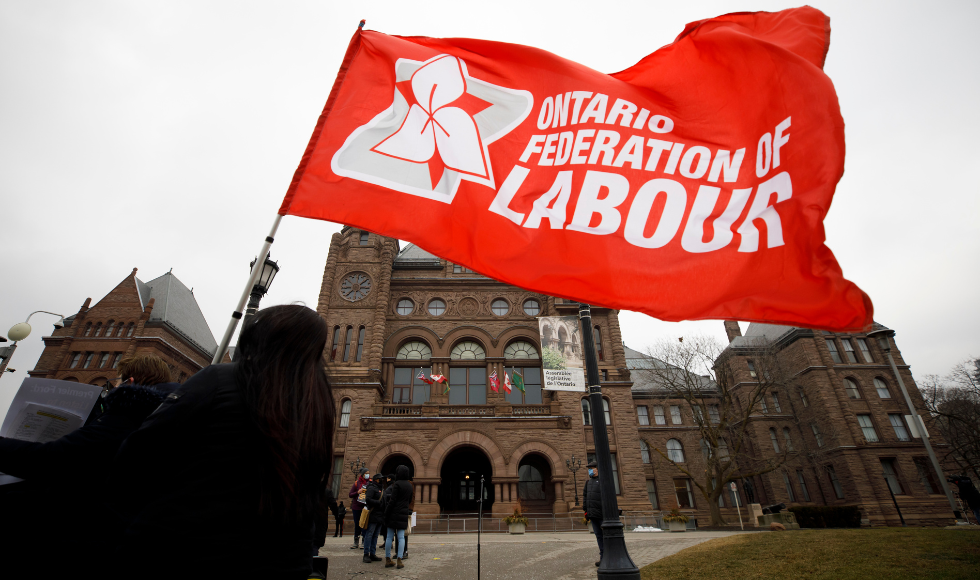 An Ontario Federation of Labour flag being held up by someone with their back to the camera. Queen's Park can be seen in the background.