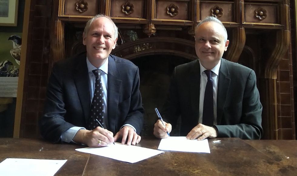 Two men in suits sitting at a table smiling at the camera while signing documents