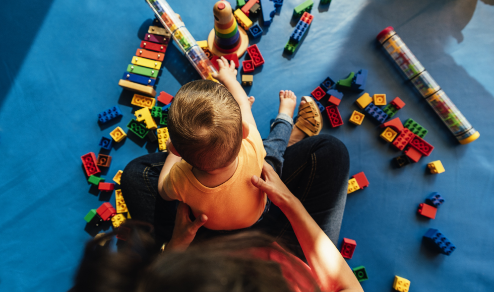 A young child sitting on the lap of an adult while playing with toys on the floor