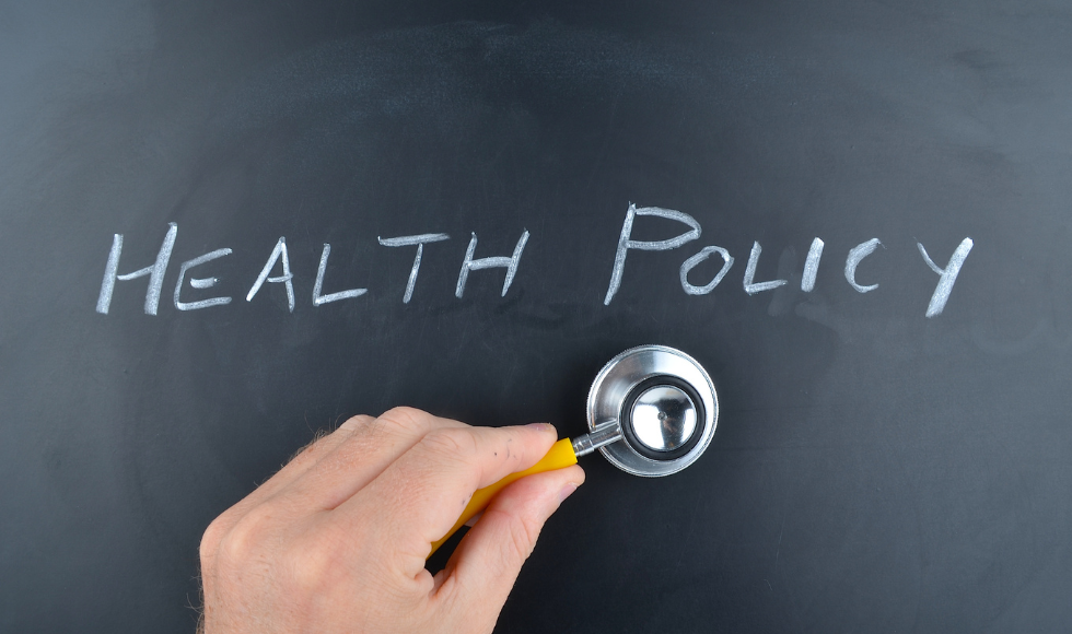 The words 'health policy' written on a chalkboard. Underneath the letters there is a hand holding up a stethoscope.