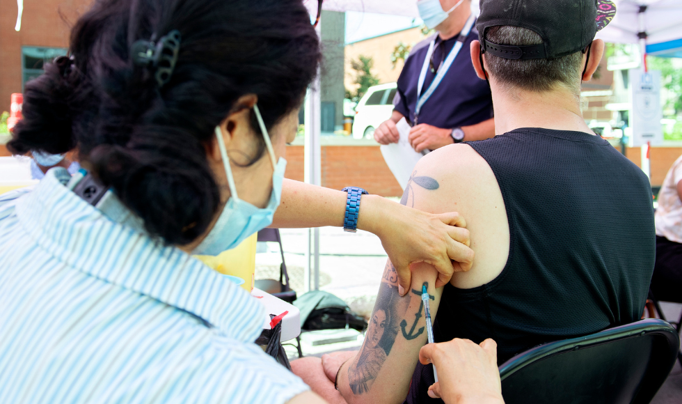 A person injecting a vaccine into the upper arm of another person. Both are masked, though the photo is taken from behind the two people and so neither face is visible.