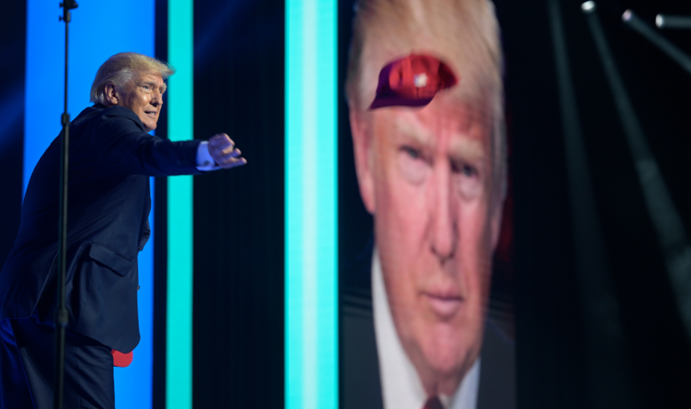 Donald Trump standing on a stage and throwing a red baseball hat. Behind him there is a large screen that has his face on it.