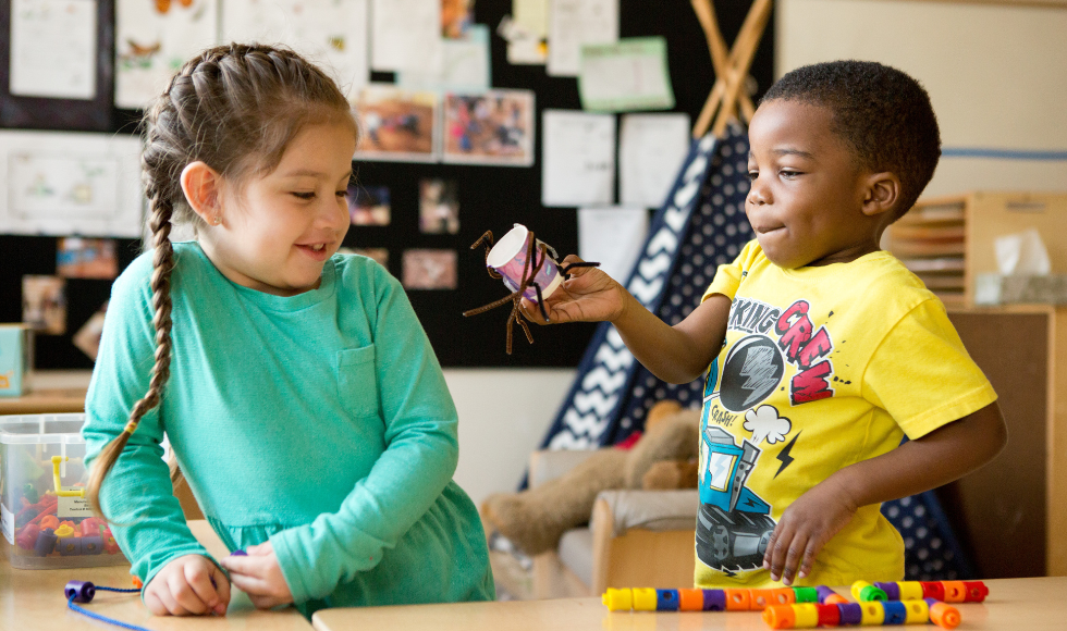 Two children smiling and looking at building blocks and crafts in a classroom.