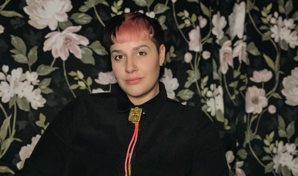 A headshot of Kaci Switzer, in a dark shirt and red bolo tie, against a dark floral background.