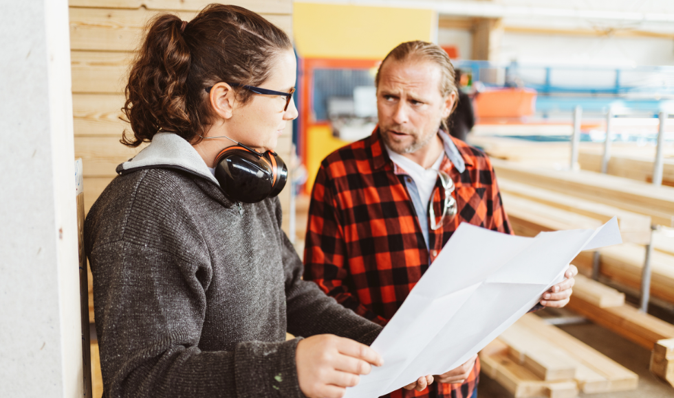 Two people holding a large piece of paper while engaged in conversation. One of the people has headphones around their neck. Piles of lumber can be seen in the background.