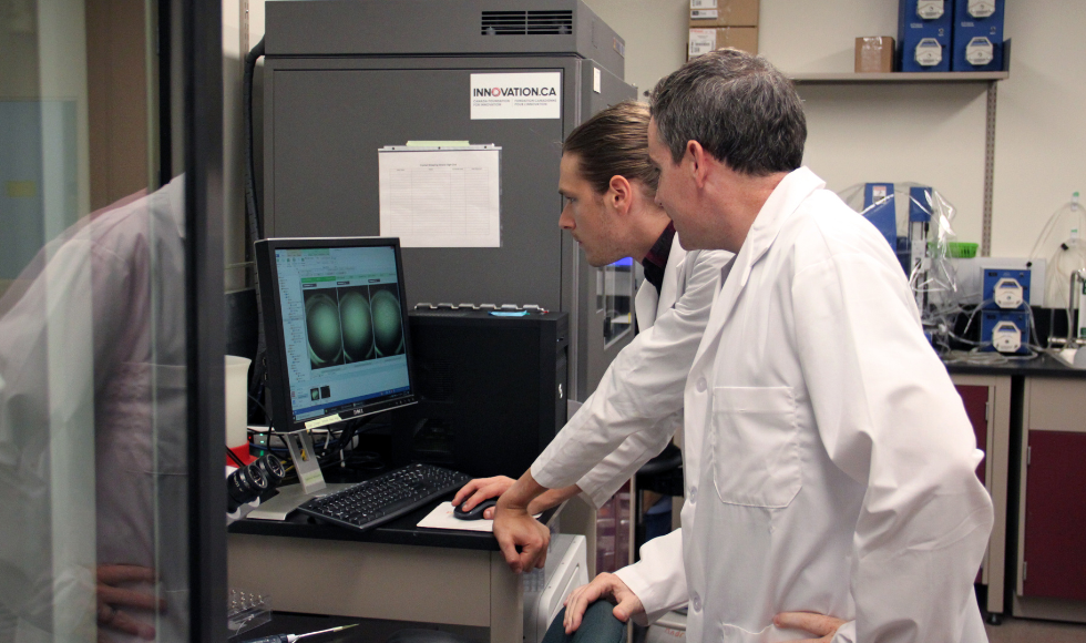 Two researchers in lab coats look at an image on a screen in a lab.