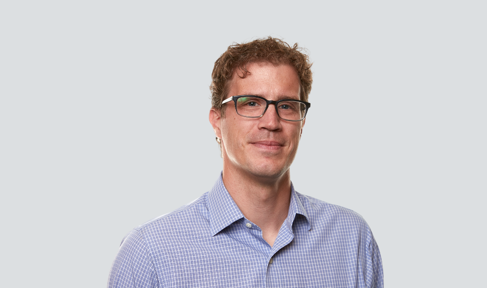 A headshot of Dominik Mertz. He is wearing a light-purple, button-up shirt and black-rimmed glasses.