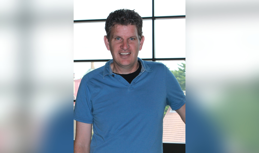 Cameron Currie standingfront of a window, wearing a blue polo shirt with a black undershirt.