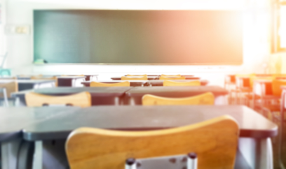 An empty classroom showing a row of desks and a green chalkboard. The photo is taken from the viewpoint of someone seated at one of the desks.