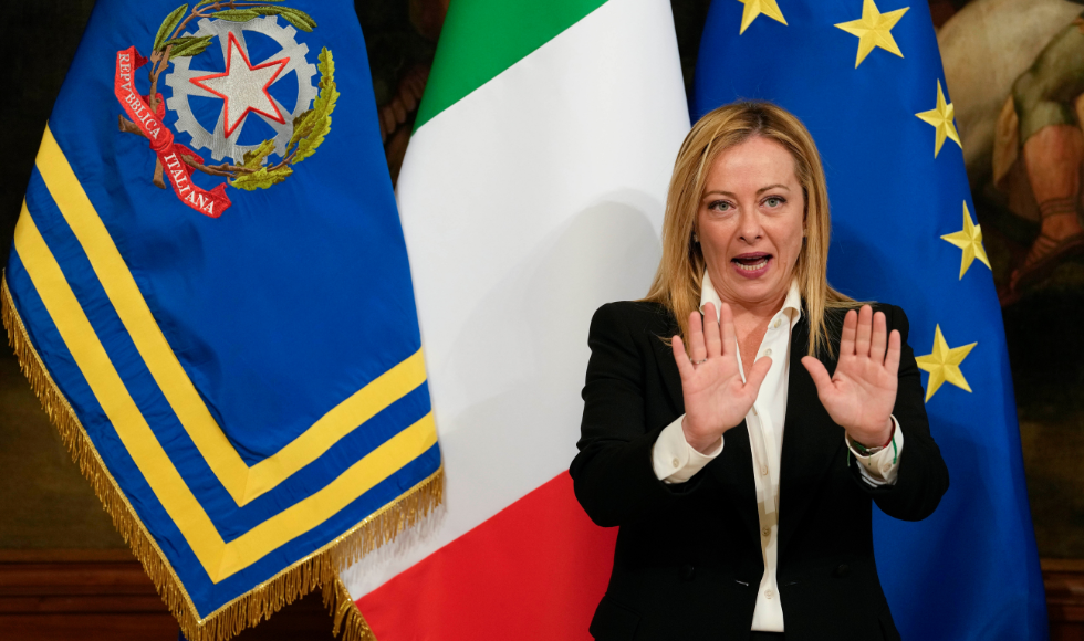 Giorgia Meloni with her hands raised and palms facing outward. She is wearing a black suit jacket, white shirt and is standing in front of three flags.