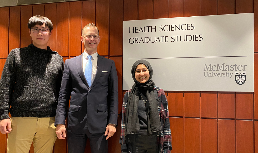 Ali Zhang, Ryan Van Lieshout and Sawayra Owais standing and smiling at the camera. Beside them is a sign that reads, ‘Health Sciences graduate studies.’