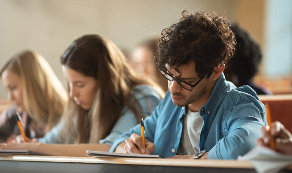 Several students with their heads bent and using pens to write. One of the students is closest to the camera and in focus, while the others are in the background and slightly out of focus.