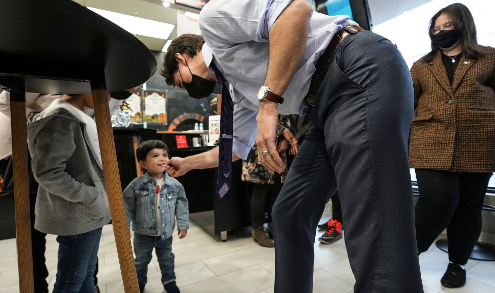 A masked Justin Trudeau bent over to speak to a young child who has what appears to be a lollypop in his mouth.