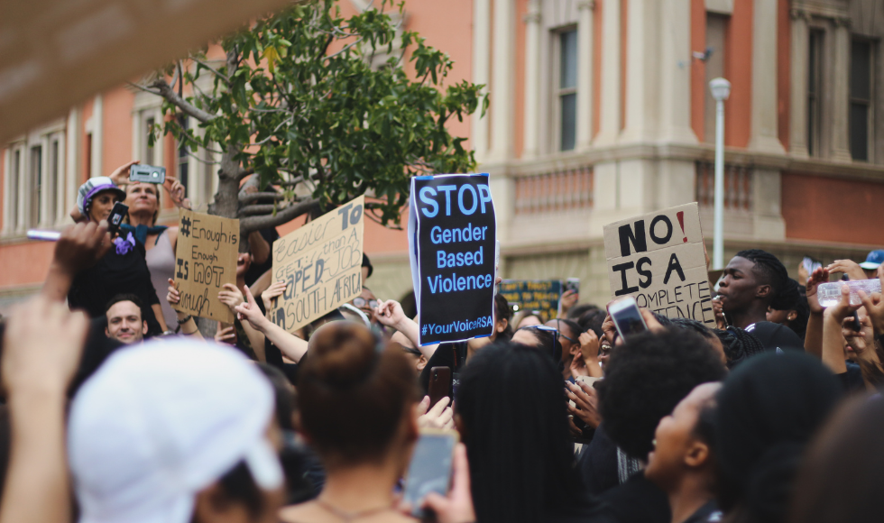 People gathered at an outdoor rally holding signs calling for a stop to gender-based violence