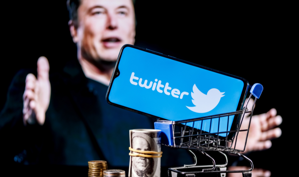 A photo illustration with a soft-focus Elon Musk in the background and a smartphone with Twitter's logo on it in the foreground, sitting in a small shopping cart, alongside stacks of cash and coins.
