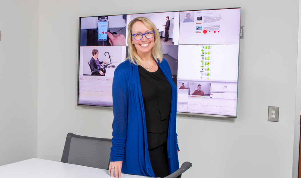 Milena head, wearing a dark top with a blue cardigan over it, standing at a table in a room with a big monitor behind her showing scenes from the MDTRC.