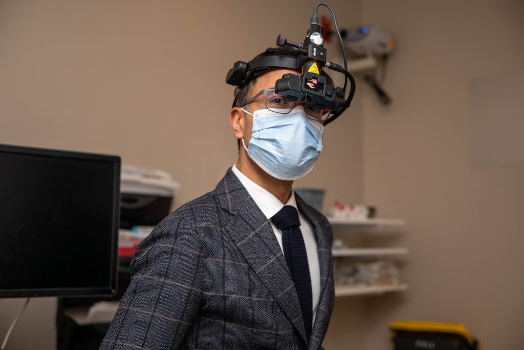Varun Chaudhary head and shoulders while he is wearing a binocular indirect ophthalmoscope on his face, as well as a medical mask, and a grey suit and tie.