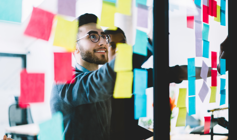 A person wearing glasses looking up at a piece of glass that has many colourful post-it notes on it.