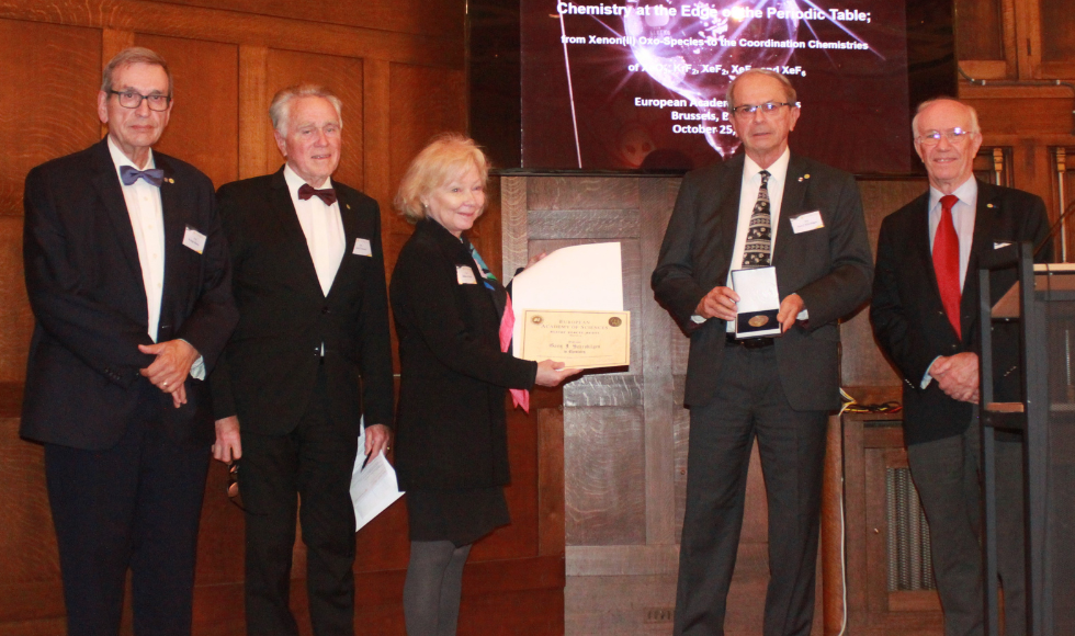 Five people standing on a stage in front of a podium. The person in the centre is holding a certificate and the person beside them is holding up an award.