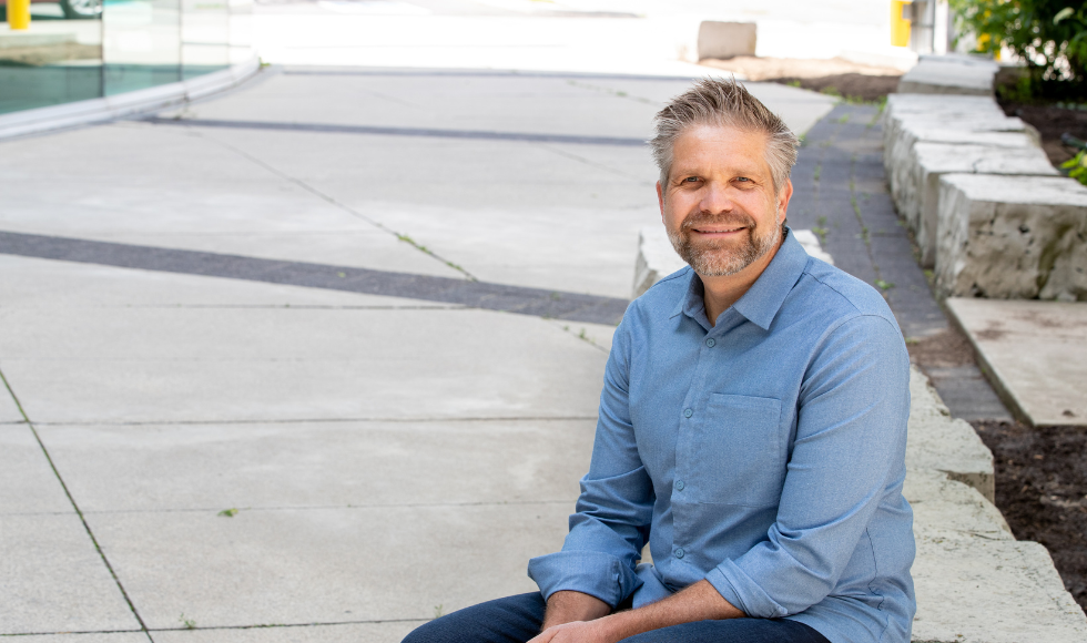 Smiling Hendrik Poinar seated outdoors wearing a blue shirt and blue pants.