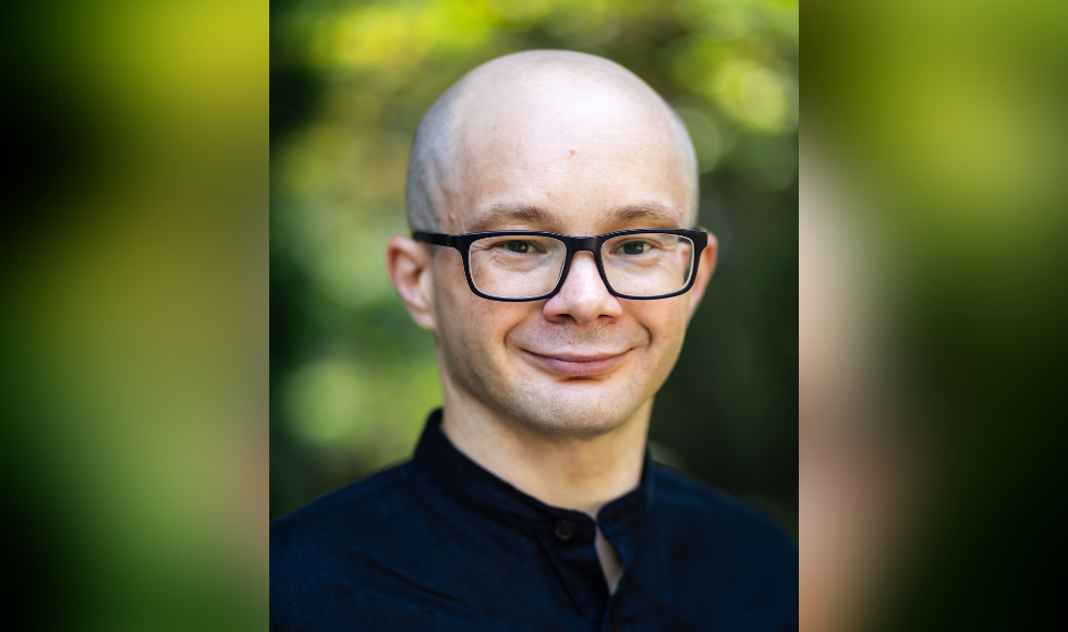 A headshot of Johannes Steizinger wearing black-rimmed glasses and a black shirt