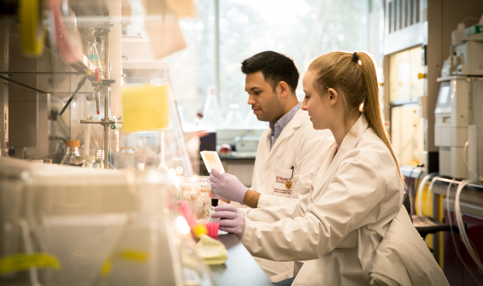 Two graduate students wearing lab coats and working in a lab full of beakers and equipment.