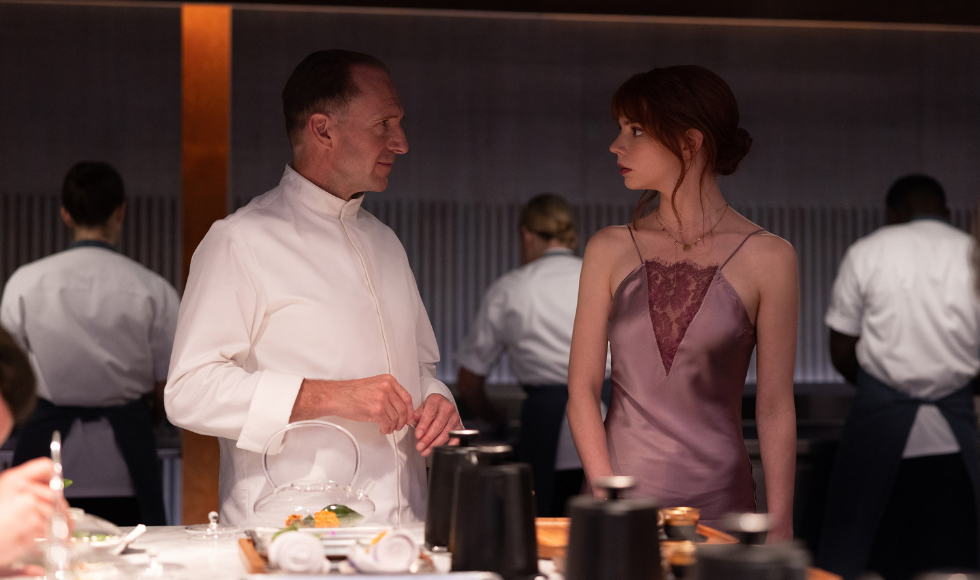 A still from The Menu showing Ralphe Fiennes and Anya Taylor Joy looking at one another near a dining table.