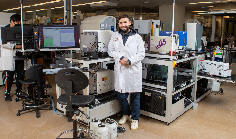 A man wearing a white lab coat standing in a laboratory setting. In the background, there is another person wearing a mask and working at a computer monitor.