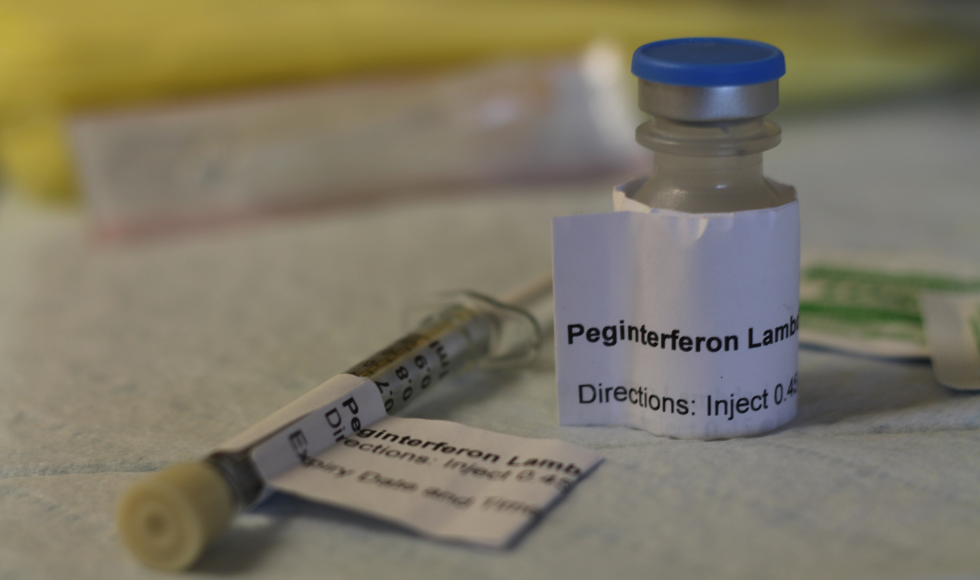 A vial and syringe sitting on a surface covered by a disposable medical cloth. Both the vial and syringe have white labels that read, ‘Peginterferon Lambda.’