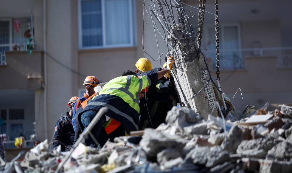 Rescue workers work together to hold up a large piece of concrete that has multiple metal rods sticking out of it. They are standing on a pile of debris and there is an apartment building behind them.