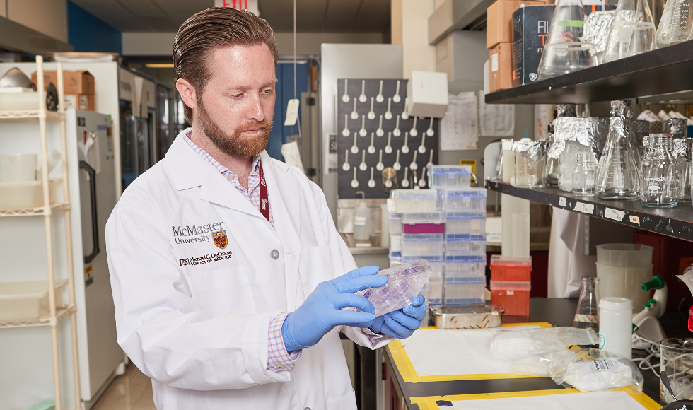 Matt Miller wearing a McMaster lab coat and latex gloves in a laboratory setting, looking at a sample