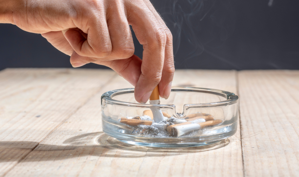 A hand stubbing out a cigarette in a transparent ashtray that is sitting on a wooden table