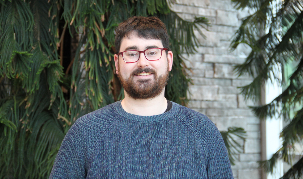 Head and shoulders of Sheridan Baker wearing a blue sweater in front of a wall and greenery.