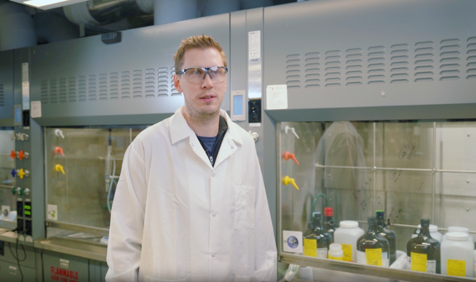 Drew Higgins in a lab coat and safety glasses standing in a lab setting.